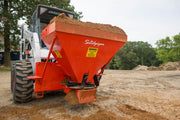 HitchDoc Sandpiper spreader heaped with damp sand on a white Bobcat skid steer, front three-quarter view showing the tapered red hopper, the warning decal and material sitting on the spreader disc below