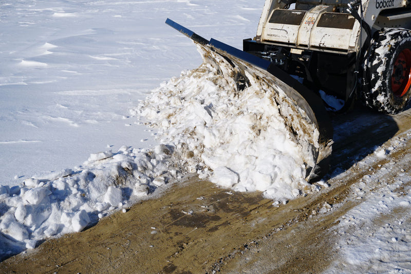 HitchDoc V-Blade angled to cast snow to one side on a Bobcat skid steer, clearing a lane down to bare pavement