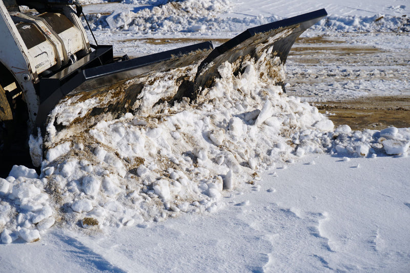 HitchDoc V-Blade loaded with snow on a skid steer, wide side view showing the mount frame and the cutting edge riding down to bare pavement