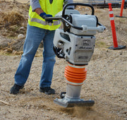 Multiquip MTX-70 tamping rammer compacting loose fill on a gravel base, an operator in a hi-viz vest steering the black tubular handle from the left, silver aluminium gearcase carrying the MQ Multiquip mark, the Mikasa script and a cast MTX-70 badge, orange protective bellows above the ram cylinder, and a bolted shoe throwing dust at the base