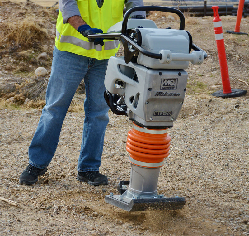 Multiquip MTX-70 tamping rammer compacting loose fill on a gravel base, an operator in a hi-viz vest steering the black tubular handle from the left, silver aluminium gearcase carrying the MQ Multiquip mark, the Mikasa script and a cast MTX-70 badge, orange protective bellows above the ram cylinder, and a bolted shoe throwing dust at the base