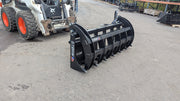 Storm HD rake and brush grapple on pavement with a skid steer behind, three-quarter view of the wide tine bank