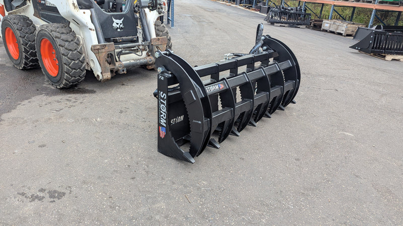 Storm HD rake and brush grapple on pavement with a skid steer behind, three-quarter view of the wide tine bank