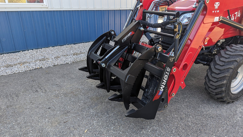 Storm rake and brush grapple on a red tractor, three-quarter view of the tine bank beside a building