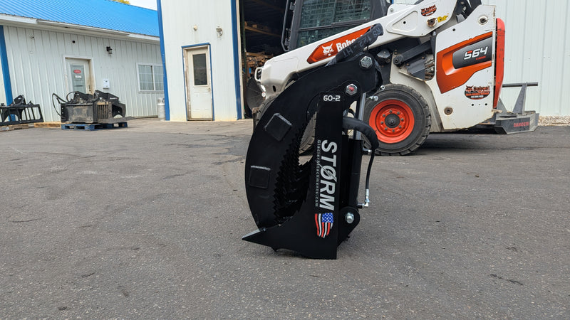 Storm two-cylinder rake and brush grapple on pavement, front view with a skid steer behind