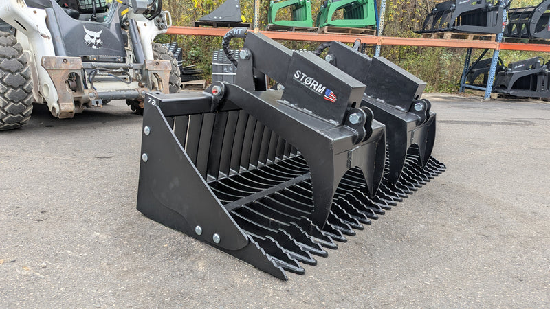 Storm rock and brush grapple on pavement with a skid steer behind, three-quarter view with the tines lowered