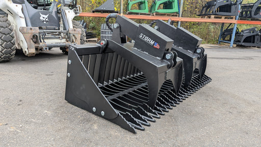Storm rock and brush grapple on pavement with a skid steer behind, three-quarter view with the tines lowered